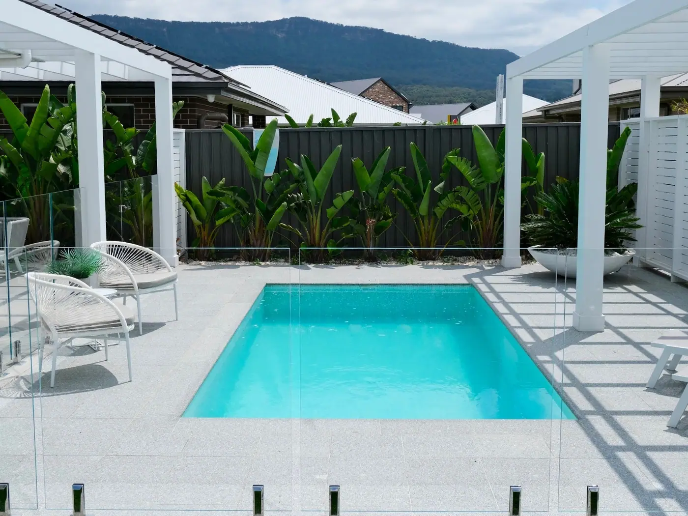 A square plunge pool in an exposed aggregate courtyard between two white pergolas, cane chairs to one side and a row of banana palms along a dark boundary fence, with hills in the distance.